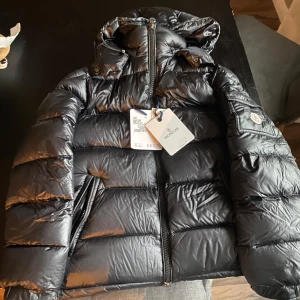 Svart Moncler Maya med huva  - Säljer en svart pufferjacka från Moncler med huva och klassisk logga på ärmen. Jackan har dragkedja framtill, två sidofickor med dragkedja och quiltad design. Perfekt för kalla dagar och riktigt snygg streetstil. Materialet är glansigt och känns lyxigt.nfc funkar👌🏼