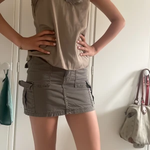 Grå cargokjol med fickor - Säljer en grå cargokjol med flera stora fickor på sidorna och bak. Kjolen är kort och har bälteshällor samt dragkedja och knapp framtill. Perfekt för dig som gillar utility-stil och vill ha en cool och praktisk look.