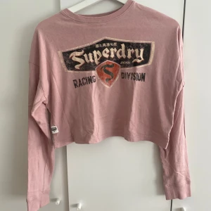 Rosa croppad långärmad Superdry tröja - Croppad långärmad tröja från Superdry i en ljusrosa nyans. Tröjan har ett fint tryck på framsidan. I mycket bra kvalitet!