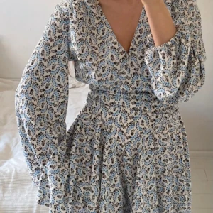 Blommig kortklänning från Maje - Supersöt kortklänning från Maje med blommigt mönster i blått och svart på vit botten. Klänningen har v-ringning, långa puffärmar och markerad midja som ger en snygg siluett. Passar M/L