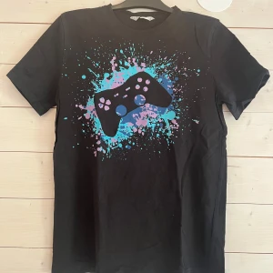 Svart gamer t-shirt från H&M - Svart t-shirt från H&M med coolt tryck av en spelkontroll och färgstänk i blått och lila på bröstet. Klassisk passform med rund hals och korta ärmar. Perfekt för dig som älskar gaming och vill sticka ut med en färgglad look.