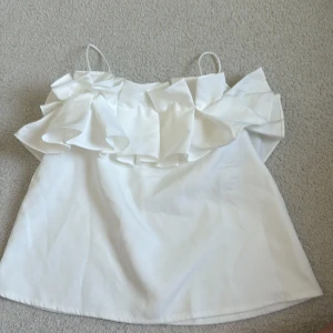 Vit volang-linnetopp med tunna band - Supersöt vit linnetopp med tunna axelband och stora volanger längs överdelen. Toppen har en luftig och lätt känsla, perfekt för varma dagar. Snygg att styla med jeans eller kjol för en fräsch look.