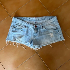 Jeansshorts med slitningar - Säljer ett par ljusblå jeansshorts som jag köpt i Spanien. Shortsen har fransiga kanter och slitna detaljer framtill och baktill. Klassisk femficksmodell med röda knappar och bälteshällor. Priset kan diskuteras!💗💗💗