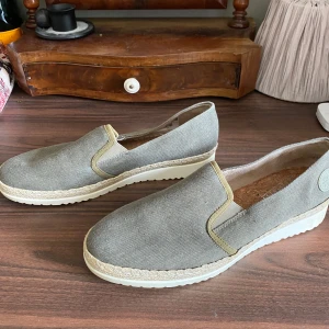 Beige slip-on loafers från Rieker - Snygga slip-on loafers från Rieker i grön canvas med ljusa detaljer och vit platt sula. Skorna har en bekväm korkinnersula och elastiska partier vid öppningen för enkel påtagning. Perfekta för en avslappnad och stilren look. Funkar för både män och kvinnor.
