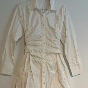 Vit skjortklänning med knappar - Stilren vit skjortklänning med lång ärm, klassisk krage och knappar hela vägen framtill. Klänningen har markerad midja med rynkade detaljer och lätt utställd kjol. Helt oanvänd.