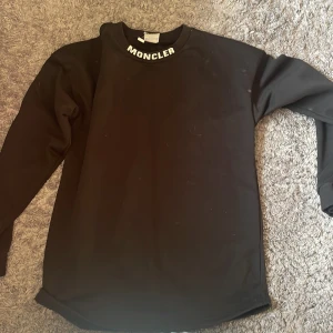 Svart långärmad Moncler tröja - Svart långärmad tröja från Moncler med vit logga tryckt både på kragen och stort över ryggen. Tröjan har en klassisk rund halsringning och är gjord i mjukt bomullsmaterial. Perfekt för dig som gillar stilrena och snygga streetwear-plagg.