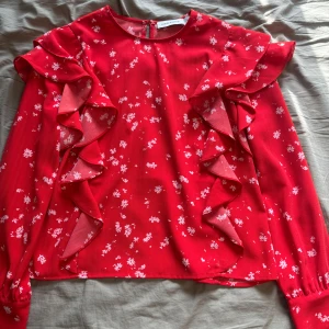 Röd blommig blus med volang Carin Wester - Supersöt röd blus från Carin Wester med vita småblommor och stora volangdetaljer längs axlar och framsida. Långärmad modell med rund halsringning och knapp i nacken. Perfekt för dig som gillar färg och lekfull stil.
