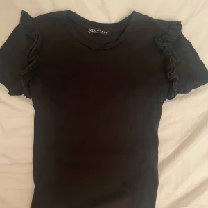Svart topp från Zara - Svart kortärmad topp från Zara med snygga volangdetaljer på axlarna. 