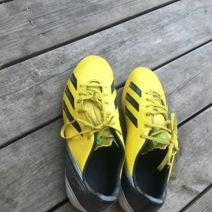 Gula Adidas fotbollsskor med dobbar - Snygga gula Adidas fotbollsskor med svarta ränder och vita sulor med gula dobbar. Skorna har snörning och är tillverkade i syntetmaterial. Perfekta för dig som vill sticka ut på planen med en sportig look.