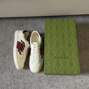 Gucci vita sneakers med orm - Snygga vita sneakers från Gucci med ikonisk röd och svart orm broderad på sidan. Skorna har klassisk snörning, rund tå och detaljer i grönt och rött på hälen. Perfekta för dig som vill sticka ut med lyxig streetstyle.