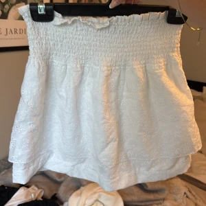 Vit broderad kortkjol från NA-KD - Supersöt vit kortkjol från NA-KD med broderade blommor och smock i midjan. Kjolen har volangkant upptill och är gjord i ett lätt bomullstyg, perfekt för varma dagar. 