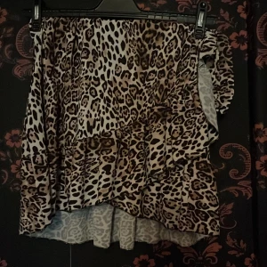 Leopardmönstrad omlottkjol från Mirell - Snygg kort omlottkjol från Mirell med leopardmönster i beige, brunt och svart. Kjolen har volangdetaljer och knytning i sidan för en cool och trendig look. Perfekt för dig som vill sticka ut med ett djurmönster.