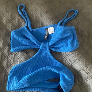 Blå topp från H&M - Snygg blå topp från H&M Divided med trendig cut out-detalj framtill och smala axelband. Materialet är strukturerat och stretchigt för en bekväm passform. Perfekt att ha till stranden.
