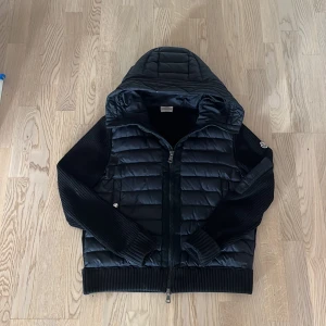 Svart Moncler cardigan  - Svart cardigan från Moncler med quiltad front och huva. Ärmarna är stickade för en schysst kontrast och jackan har dragkedja framtill. Perfekt för kyliga dagar när du vill ha både stil och värme. Klassisk Moncler-logga på ärmen.