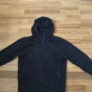 Svart hoodiejacka från C.P. Company - Svart hoodiejacka från C.P. Company med hel dragkedja, två fickor med dragkedja och ikoniska goggles i huvan. Jackan har snörning i huvan och ribbade muddar. Perfekt för dig som gillar streetwear och vill sticka ut med en unik detalj är sliten i höger ärm som man ser på tredje bilden och är solblekt annars inget fel på den självklart äkta priset är inte hugget i sten skriv om ni har frågor