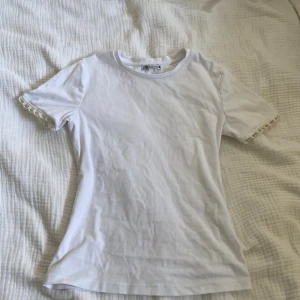 Vit topp med pärldetaljer från Zara - Supersöt vit topp från Zara med korta ärmar och unika pärldetaljer längst ut på ärmsluten. Enkel och stilren design i mjuk bomull, perfekt för dig som gillar att sticka ut med små detaljer. Snygg passform som sitter bra på kroppen.