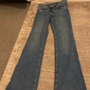Blå bootcut jeans med hög midja - Säljer ett par klassiska blå jeans med bootcut-modell och hög midja. Jeansen har fem fickor, dragkedja och knapp framtill samt en liten sliten detalj vid ena fickan. Perfekta för dig som gillar retrovibbar och en snygg siluett.