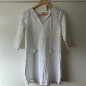 Vit broderad kortklänning från Indiska - Superfin vit kortklänning från Indiska i ett super fint broderade hålmönster med super söta knytband med tofsar på. Använd ett mycket fåtal gånger. Storlek 36