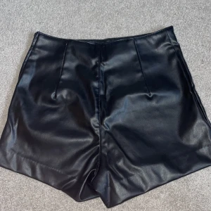 Svarta skinnshorts - Snygga svarta shorts i skinnimitation med hög midja. Från H&M och är i storlek 38. Har använt 1 gång. Shortsen har en dragkedja på sidan samt en knapp bredvid dragkedjan. Små i storlek 