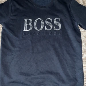 Svart tröja från Hugo Boss - Snygg svart tröja från Hugo Boss med stort BOSS-tryck på framsidan. Perfekt för en stilren look. Tröjan har korta ärmar och rund halsringning.