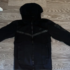 Svart jacka från Nike - Säljer en stilren svart jacka från Nike med huva och dragkedja. Jackan har diskreta detaljer och en logotyp på bröstet. Perfekt för en sportig look.