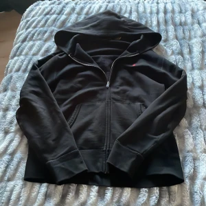 Svart hoodie från Polo Ralph Lauren - Säljer en stilren svart hoodie från Polo Ralph Lauren med dragkedja och huva. Den har en liten röd logga på bröstet och praktiska fickor framtill. Perfekt för en avslappnad look.
