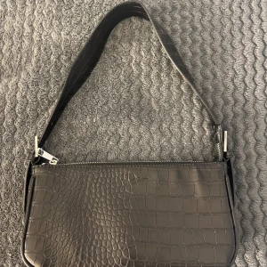 Svart axelväska  - Snygg svart axelväska med krokodilmönster och dragkedja. Perfekt för att ge din outfit en stilfull touch. Väskan har en praktisk storlek och ett elegant utseende.