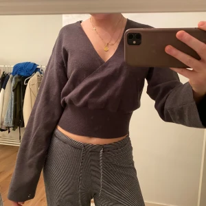 Mörkgrå omlottopp - Säljer en stilren mörkgrå omlottopp med långa ärmar. Toppen är croppad och har en v-ringad design som ger en elegant touch. Perfekt för att matcha med både jeans och kjol. Köpt på bikbok och endast testad!
