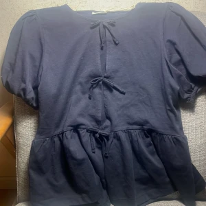 Mörkblå topp från Gina Tricot - Säljer en söt mörkblå topp från Gina Tricot med korta puffärmar och knytband framtill. Toppen har en volangdetalj vid nederkanten som ger en fin siluett. Perfekt för en stilren och avslappnad look.