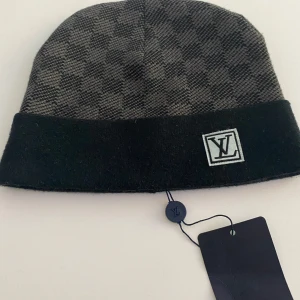 Rutig mössa från Louis Vuitton - Snygg och stilren mössa från Louis Vuitton i grått och svart med ett diskret rutigt mönster. Mössan har en uppvikt kant och en broderad logotyp framtill. Perfekt för att hålla värmen med stil.