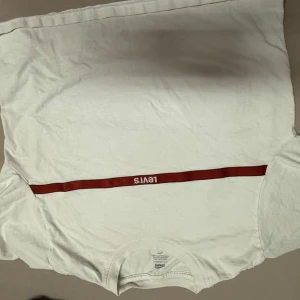 Vit t-shirt från Levi's - Säljer en klassisk vit t-shirt från Levi's med en röd rand och logga över bröstet. Perfekt för en avslappnad stil. Tillverkad i mjukt material för extra komfort.