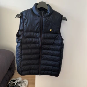 Svart dunväst från Lyle & Scott - Säljer en stilren svart dunväst från Lyle & Scott. Västen har en dragkedja framtill och ett litet gult emblem på bröstet. Perfekt för kyligare dagar när du vill hålla värmen utan att kompromissa med stilen.