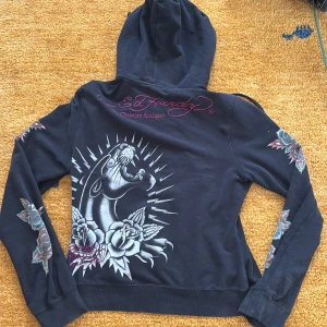 Svart huvtröja från Ed Hardy - Snygg svart huvtröja från Ed Hardy med ett stort tryck av en panter och rosor på ryggen. Tröjan har långa ärmar med matchande tryck och en dragkedja framtill. Perfekt för en cool och avslappnad stil.