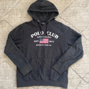 Mörkgrå hoodie från Polo Club - Säljer en snygg mörkgrå hoodie från Polo Club med tryck framtill och en amerikansk flagga. Hoodien har en klassisk känguruficka och justerbar huva. Perfekt för en avslappnad stil.