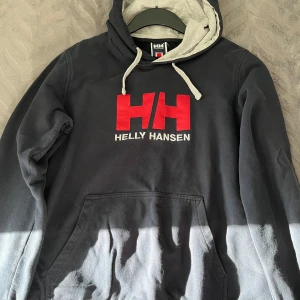 Svart hoodie från Helly Hansen - Säljer en svart hoodie från Helly Hansen med röd logga på bröstet. Den har en grå insida i huvan och en praktisk magficka. Perfekt för kyliga dagar.