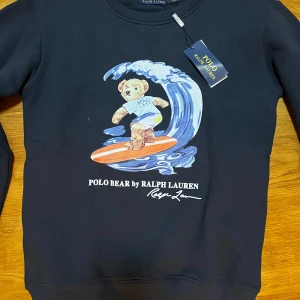 Mörkblå sweatshirt med Polo Bear från Ralph Lauren - Säljer en mörkblå sweatshirt från Ralph Lauren med ett coolt tryck av Polo Bear som surfar på en våg. Tröjan har långa ärmar och rund halsringning. Perfekt för en avslappnad stil med en touch av humor.