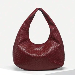 Vinröd axelväska - Snygg vinröd axelväska med flätat mönster. Väskan har en unik form och är perfekt för att ge din outfit en extra touch. Tillverkad i syntetmaterial som ger en lyxig känsla. Nyskick 