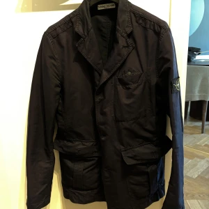 Mörkblå jacka från Stone Island - Säljer en stilren mörkblå jacka från Stone Island. Jackan har en klassisk design med knappar och flera praktiska fickor. Perfekt för höst och vår.