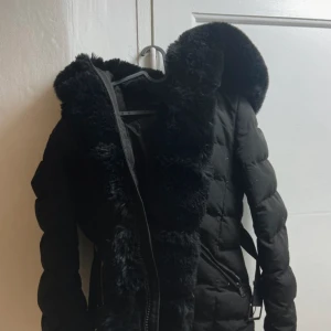 Svart dunjacka med pälskrage - Snygg svart dunjacka med fluffig pälskrage som kan dras av och bälte i midjan. Köpt för 1500kr💕💕Finns ett litet hår bakom vid axeln som går att sy.