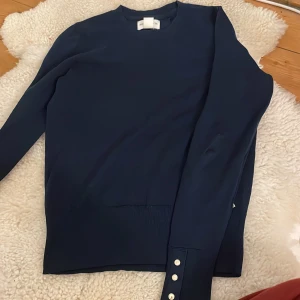 Mörkblå tröja med knappar - Säljer en stilren mörkblå tröja med långa ärmar och dekorativa knappar vid ärmsluten. Tröjan har en ribbad struktur vid muddar och nederkant, vilket ger en snygg detalj. Perfekt för en elegant look.