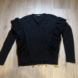 Svart tröja med volangdetaljer från Mango - Snygg svart tröja från Mango med volangdetaljer längs sidorna. Tröjan har en v-ringad hals och långa ärmar, perfekt för en stilren look. Passar bra till både jeans och kjol.