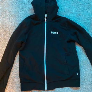 Svart hoodie från Boss - Säljer en stilren svart hoodie från Boss med dragkedja och huva. Tröjan har långa ärmar och en vit logotyp på bröstet. Perfekt för en avslappnad look.