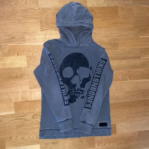 Grå hoodie med dödskalle från Skullsnbones - Snygg grå hoodie från Skullsnbones med en stor svart dödskalle på framsidan och text längs ärmarna. Perfekt för en avslappnad stil med en edgy touch. Huvan ger extra komfort och stil. Storlek Xs/146/152