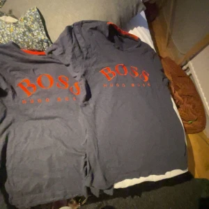 2 t-shirt från Hugo Boss - Säljer två svart t-shirt från Hugo Boss med röd logga på bröstet. Perfekt för en stilren och avslappnad look. T-shirten har korta ärmar och är tillverkad i ett mjukt material.