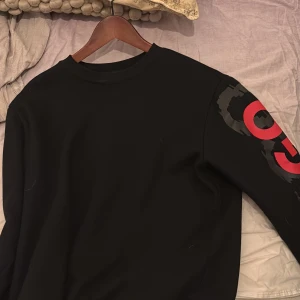 Svart tröja från Hugo - Snygg svart tröja från Hugo med röd logga på ärmen. Perfekt för en stilren look. Tröjan har långa ärmar och en klassisk passform.