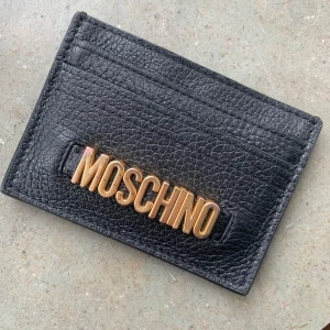 Svart plånbok från Moschino - Jättesöt liten och behändig korthållare i svart läder från Moschino. Givetvis äkta. Köpt på Raglady by Tara för ca 1 år sedan. Knappt använd!   Nypris 2.629kr
