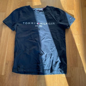 Svart t-shirt från Tommy Hilfiger - Snygg svart t-shirt från Tommy Hilfiger med tryck på bröstet. Perfekt för en avslappnad stil. Klassisk rund hals och korta ärmar.