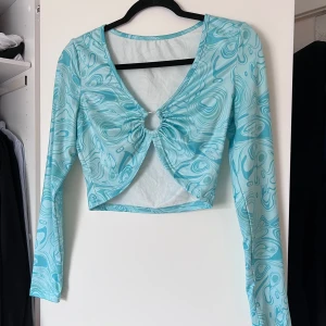 Turkos mönstrad topp - Säljer en snygg turkos topp med ett unikt mönster och långärmad design. Toppen har en snygg detalj framtill med en ring som ger en extra touch. Perfekt för att sticka ut i mängden! Aldrig använd 🩵 Önskar du veta exakta mått? Tveka inte att höra av dig!