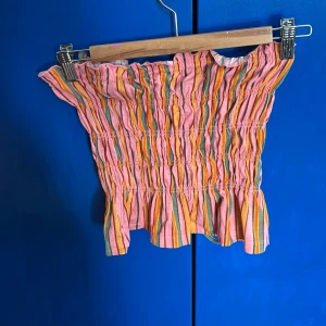 Randig tubtopp i rosa och orange - Säljer en färgglad randig tubtopp i rosa, orange och grönt. Toppen har en smockad design med volangdetaljer upptill och nedtill. Lappen är bortklippt från innan jag köpte den, aldrig använt den själv. Det kommer med band på toppen också🧡🩷💛