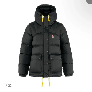 Svart dunjacka från Fjällräven - Säljer en svart dunjacka från Fjällräven med huva och gula detaljer. Jackan har dragkedja och knappar framtill samt två stora fickor. Perfekt för kalla dagar. Jackan är i storlek xs och tjej modell 